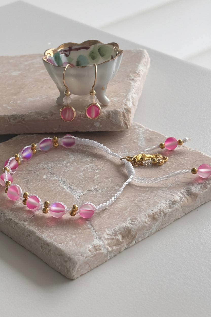 A handmade pink mermaid glass macrame bracelet with brass spacer beads and a gold mermaid charm, displayed on a pink stone slab alongside matching pink cat's eye drop earrings with gold hooks in a small abalone dish. Each item sold separately.