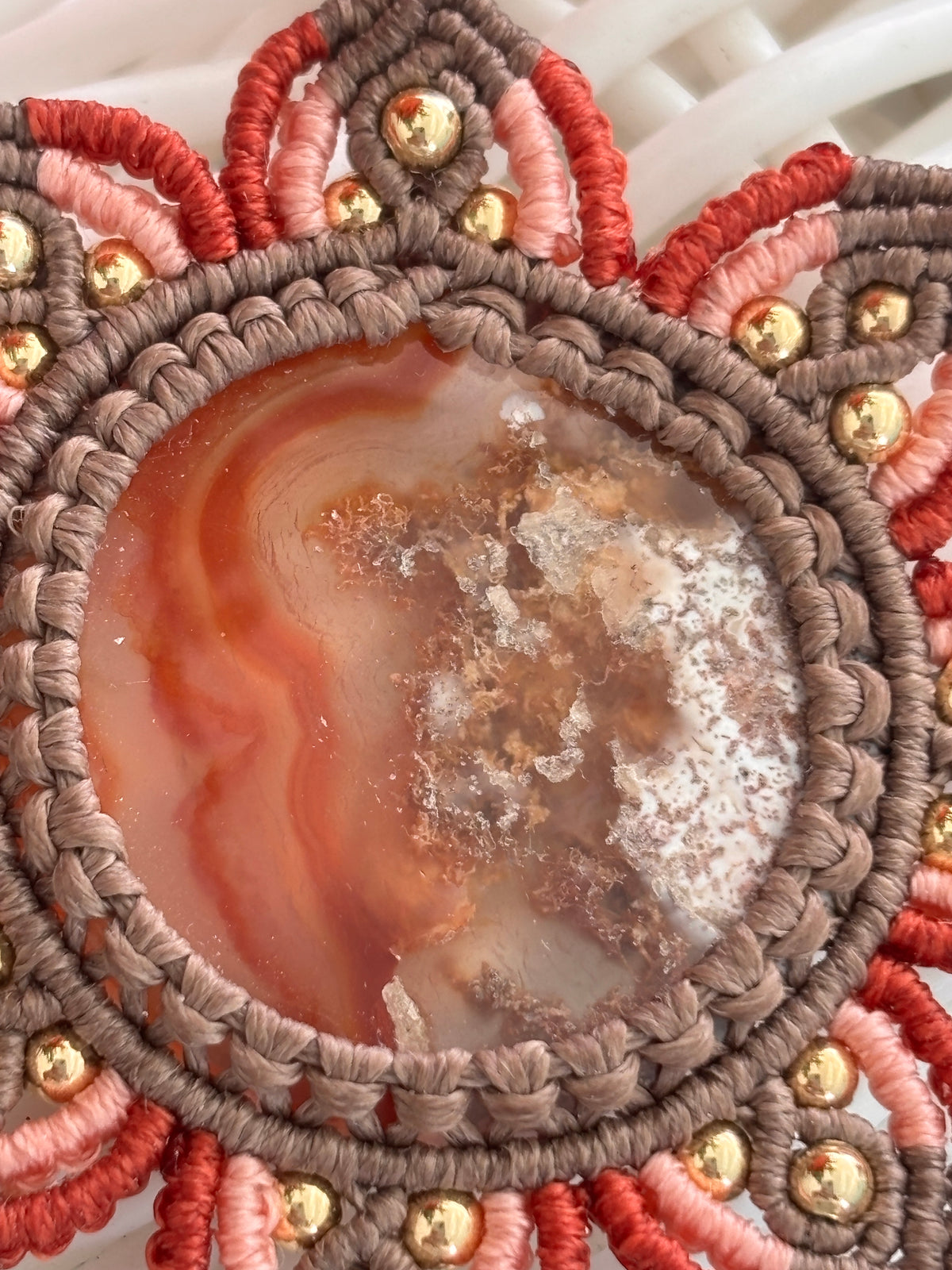 A close-up of a handmade macrame necklace pendant featuring a central round Carnelian Agate stone with swirling orange, brown, and white patterns, surrounded by a thick brown braided border and radiating rust, peach, and brown details.