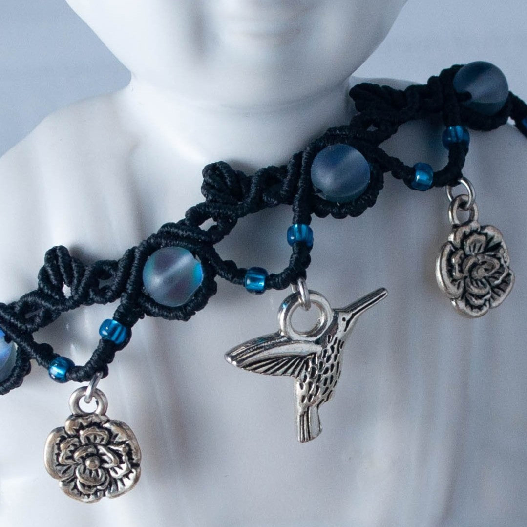 A close-up of a black macrame anklet displayed on a white budha figure. The anklet features frosted iridescent blue mermaid glass beads, small blue seed beads, a silver-tone hummingbird charm, and two silver floral charms.
