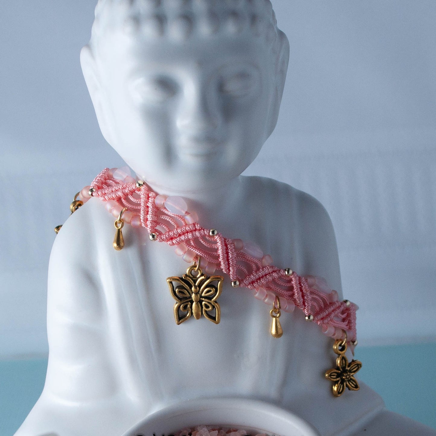 A coral macrame beaded anklet with gold-tone butterfly and waterdrop charms draped over a white Buddha statue against a soft light blue background.