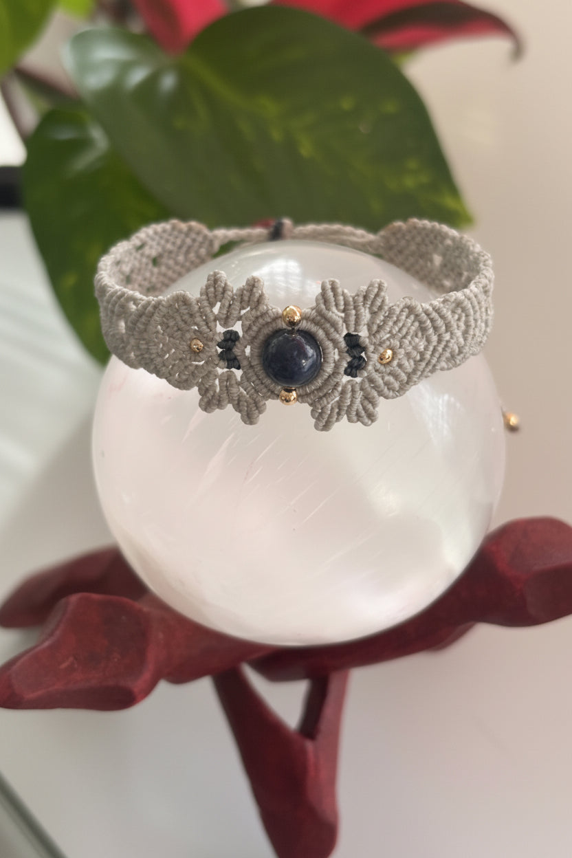 A handmade gray macrame bracelet with a central lapis lazuli gemstone and small brass accent beads, displayed on a white crystal sphere.