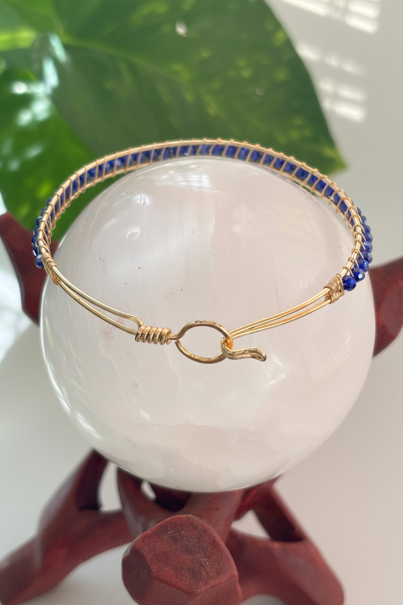 A handmade brass wire bangle bracelet with a blue faceted crystal beads, displayed on a white stone sphere.