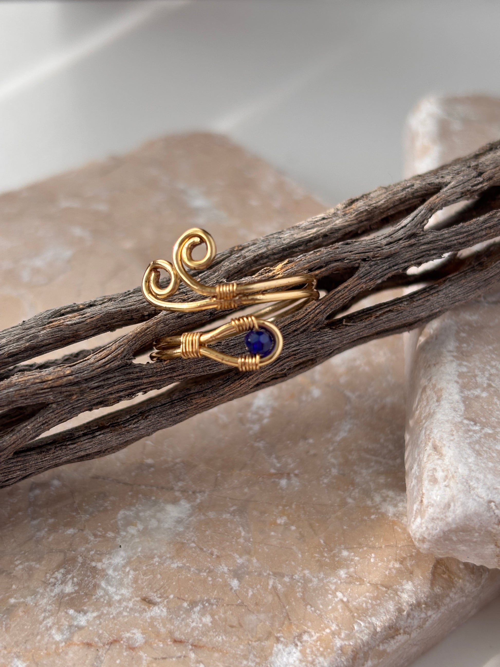 A handcrafted gold-tone wire ring with a double spiral flourish and a 3mm deep blue faceted crystal bead wire-wrapped at the base, resting on weathered driftwood across a natural stone slab against a white background.