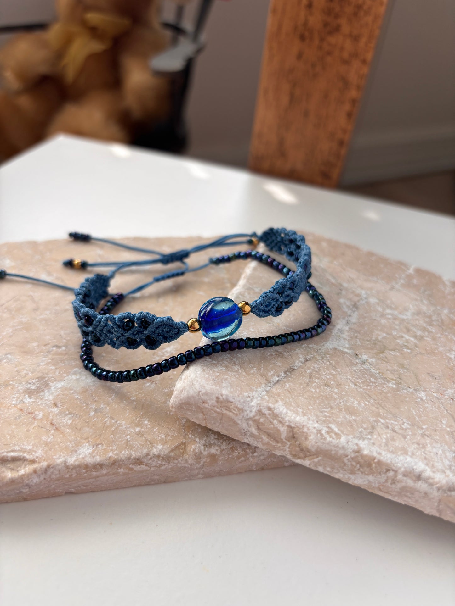 A handcrafted dark blue iridescent seed bead bracelet stacked with a medium blue macrame bracelet featuring an aqua glass disc bead and gold-tone accents, displayed on overlapping stone slabs against a white background. Items sold separately.