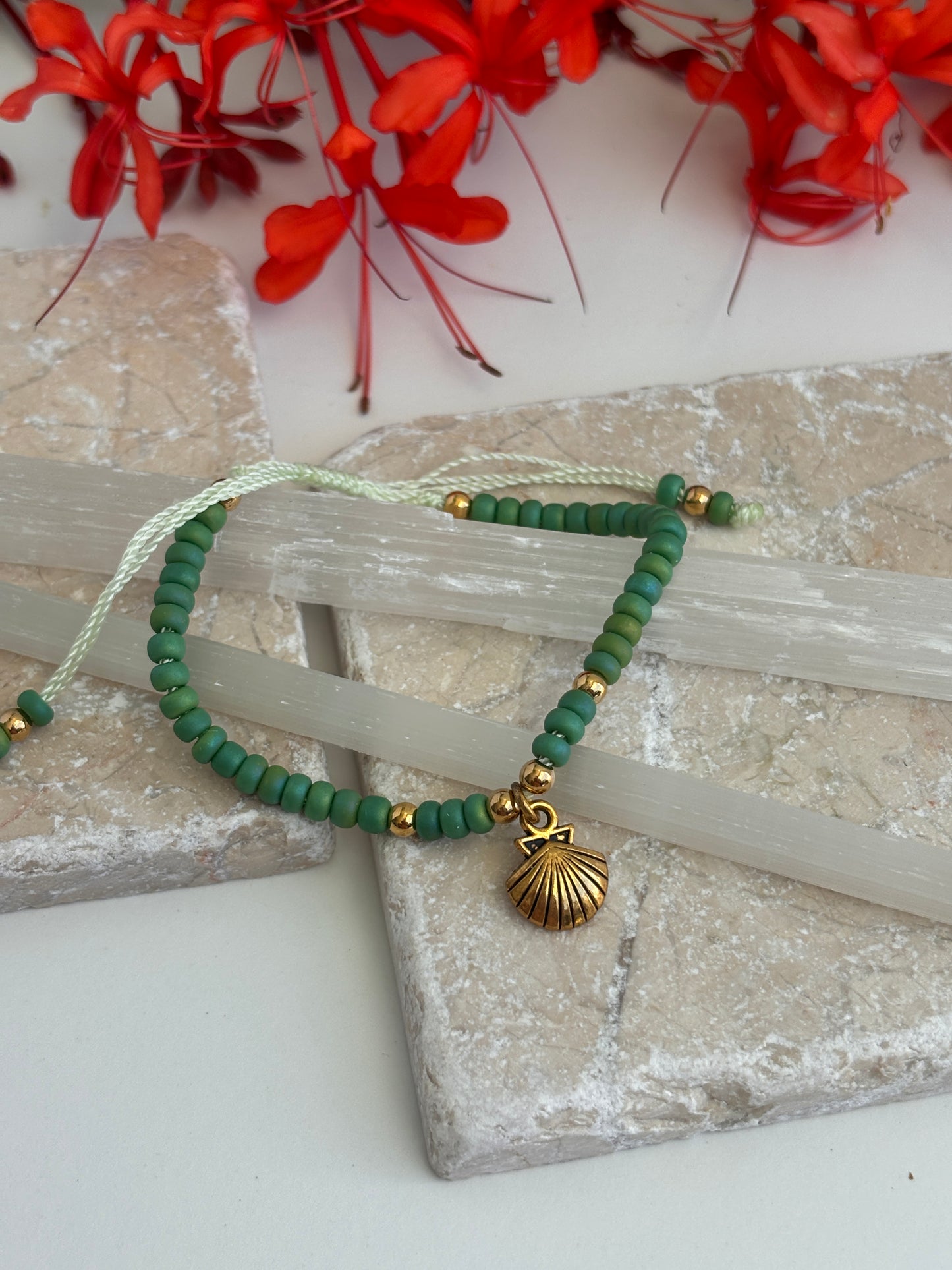 A green beaded bracelet with mint green cord, small brass spacer beads, and a brass shell charm, displayed on a stone slab with red flowers in the background.