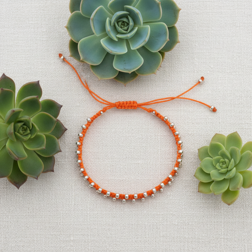 An orange macrame bracelet with small galvanized silver seed beads, laid flat on a textured off-white fabric background and surrounded by three green succulent plants.