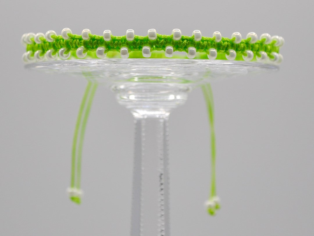 A lime green macrame bracelet or anklet with pearlescent white seed beads displayed on a clear glass stand against a light gray background.