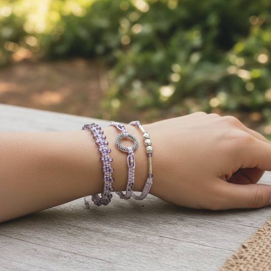 A close-up of a person's wrist wearing three delicate lavender macrame bracelets with lavender seed beads and various shaped metal accent beads, resting on a weathered wooden surface with a soft-focus green garden background. 