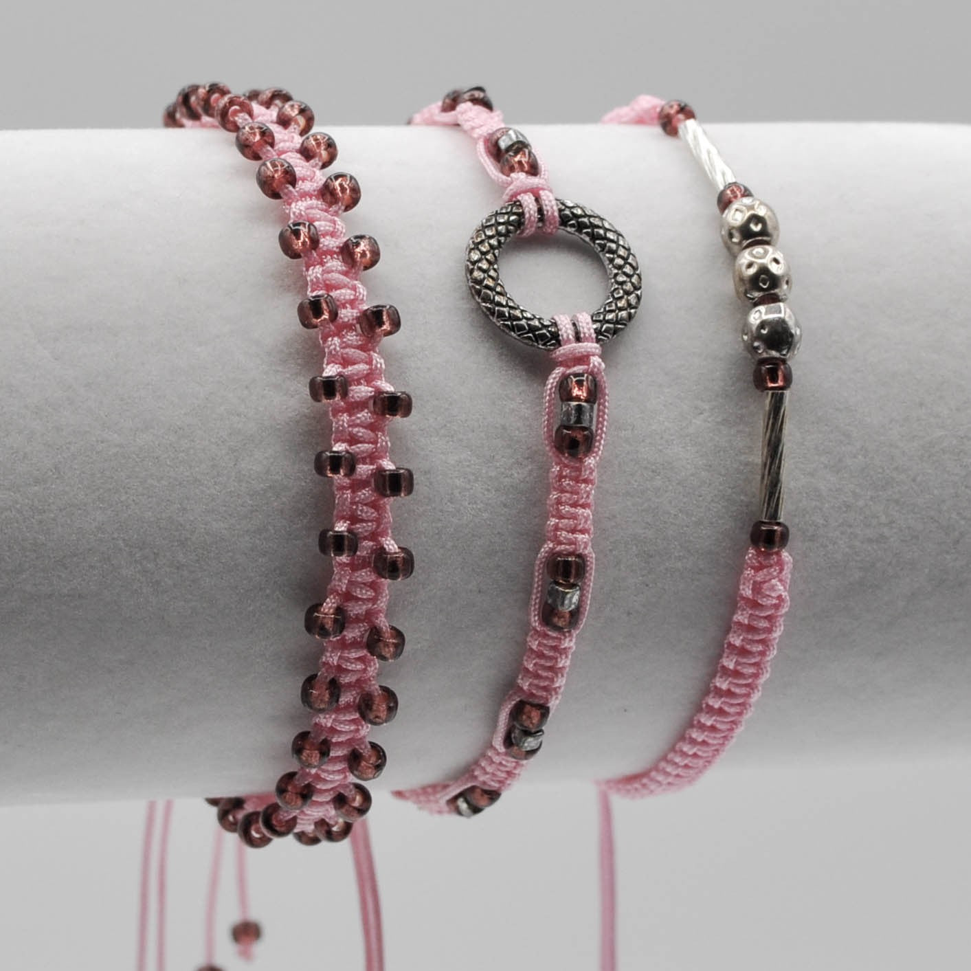 Three pink macrame bracelets with cranberry seed beads and various shaped metal beads displayed on a white cylindrical jewelry stand against a light gray background.
