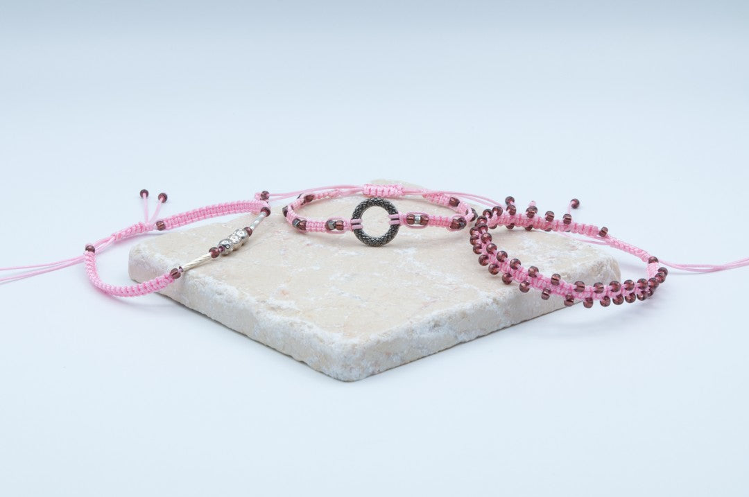 Three pink macrame bracelets with cranberry seed beads and various shaped metal accents, displayed on a light beige stone slab against a white background.
