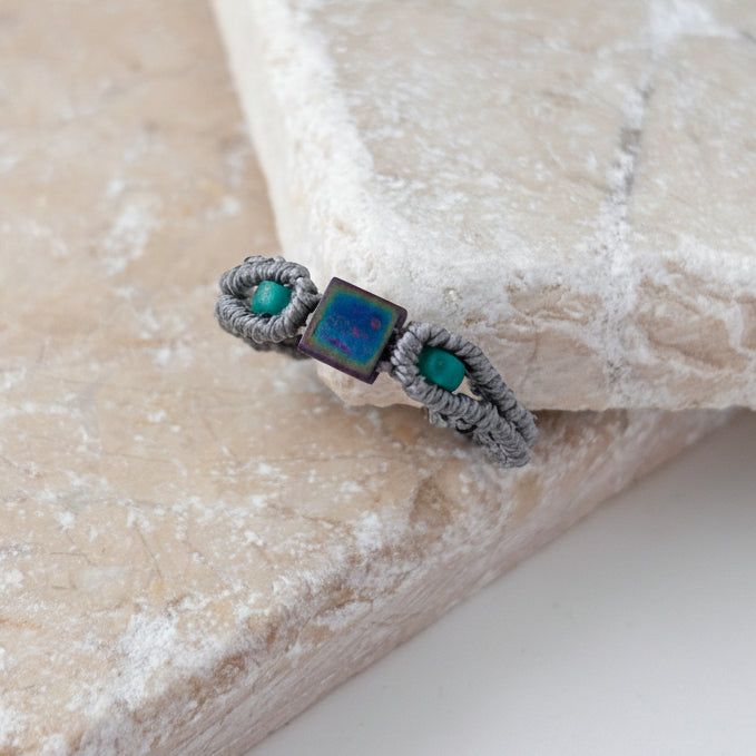 A close-up of a handmade minimalist gray macrame ring with an iridescent blue-purple square Miyuki Tila bead at the center and two teal seed bead accents, resting on the edge of a pink stone surface against a white background.
