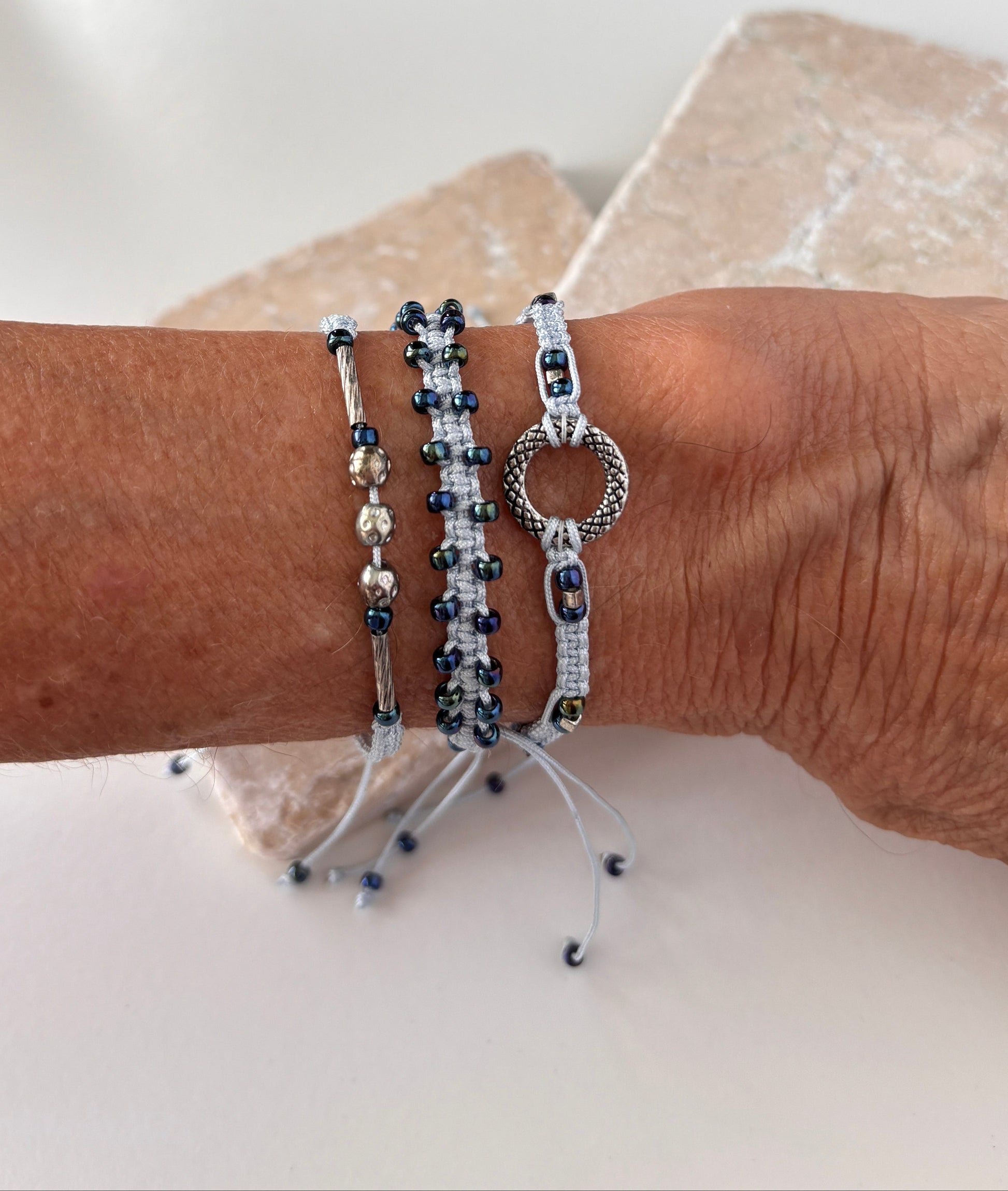 Three light blue macrame bracelets stacked on a wrist against a white stone background — one with a silver-tone open ring centerpiece, one with dark metallic blue seed beads woven along the edges, and one with a silver-tone tube bead and silver-tone ball accent beads on light blue nylon cord.