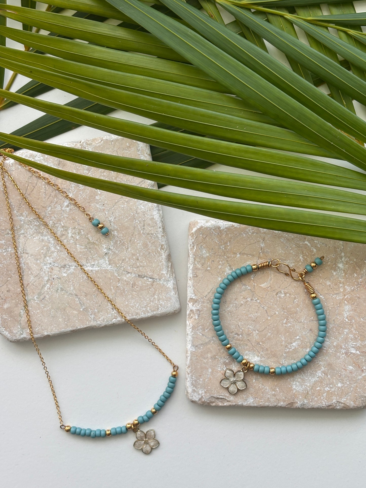 Brass jewelry set with turquoise blue seed beads and white enamel flower charm, including a necklace and bracelet, styled on a white surface with natural stone blocks and green palm leaves.  Each item sold separately.