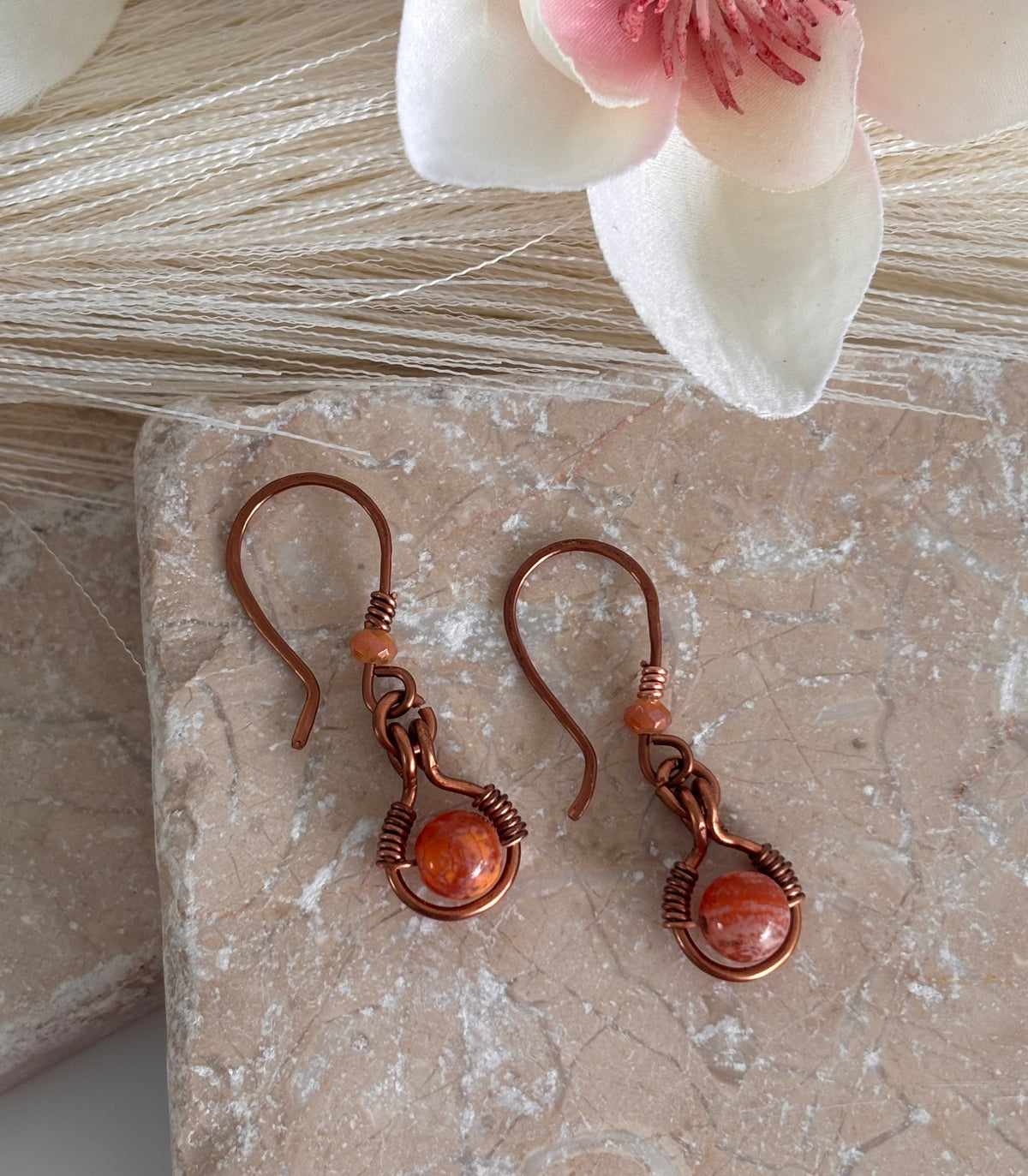 Handmade Ancient Cellar agate drop earrings with antiqued copper wire halo and iridescent orange crystal bead, displayed on travertine tile with white magnolia bloom and pampas grass — artisan boho earrings.