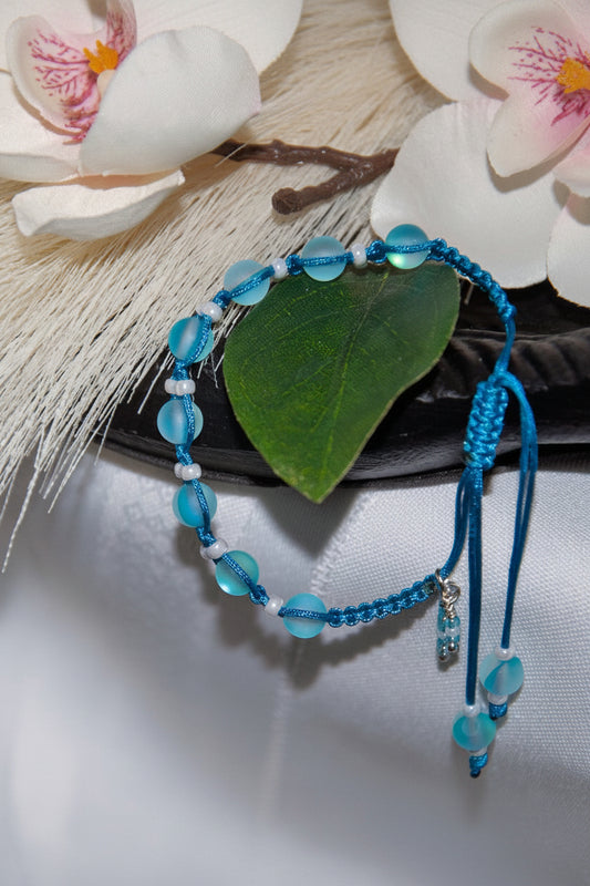 A beaded macrame bracelet with frosted aqua round beads, white pearl seed bead spacer beads, and a small seed bead dangle charm, styled on a white fabric surface with a green leaf and white flowers in the background.