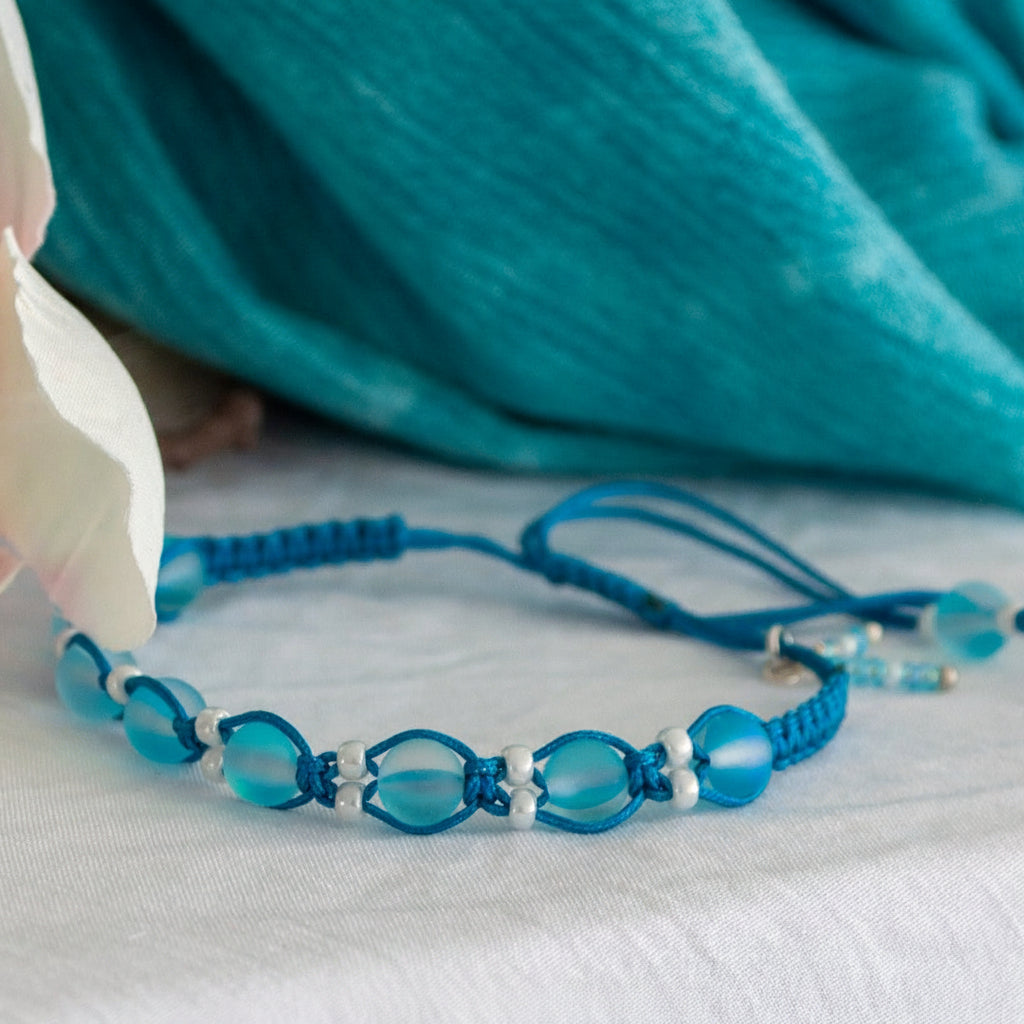 A beaded macrame bracelet with frosted aqua round beads and white pearl seed bead spacer beads, resting on a white fabric surface with a teal fabric backdrop and a white flower petal.