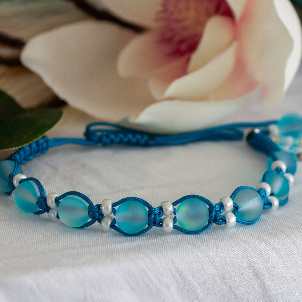 A handmade beaded macrame bracelet with frosted aqua round beads and teal cord, accented with small white pearl seed beads, displayed on a white fabric surface with a blurred white flower in the background.