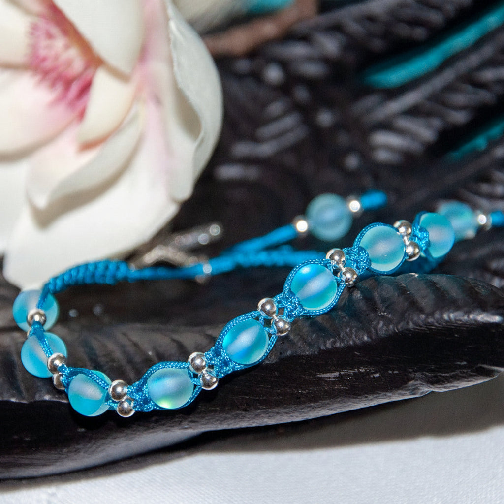 A handmade teal blue beaded macrame bracelet with teal blue frosted mermaid glass round beads, and silver-tone spacer beads, displayed on a dark textured surface with a white flower in the background.