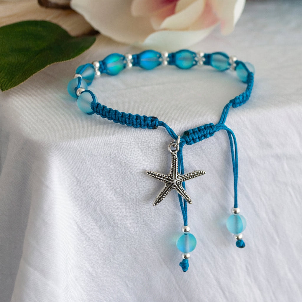 A teal blue macrame bracelet with round frosted teal blue mermaid glass beads, silver-tone spacer beads, and a silver-tone starfish charm, and dangling bead accents, displayed on a white fabric surface with a soft blurred flower in the background.