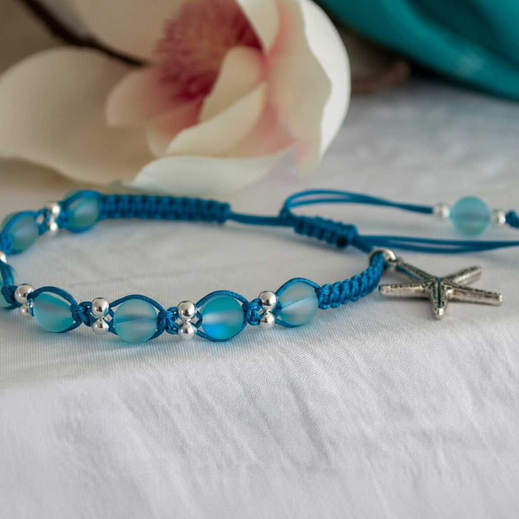 A teal blue beaded macrame bracelet with frosted teal blue mermaid glass beads, small silver-tone spacer beads, and a silver-tone starfish charm, displayed on a white textured fabric surface with a blurred flower in the background.