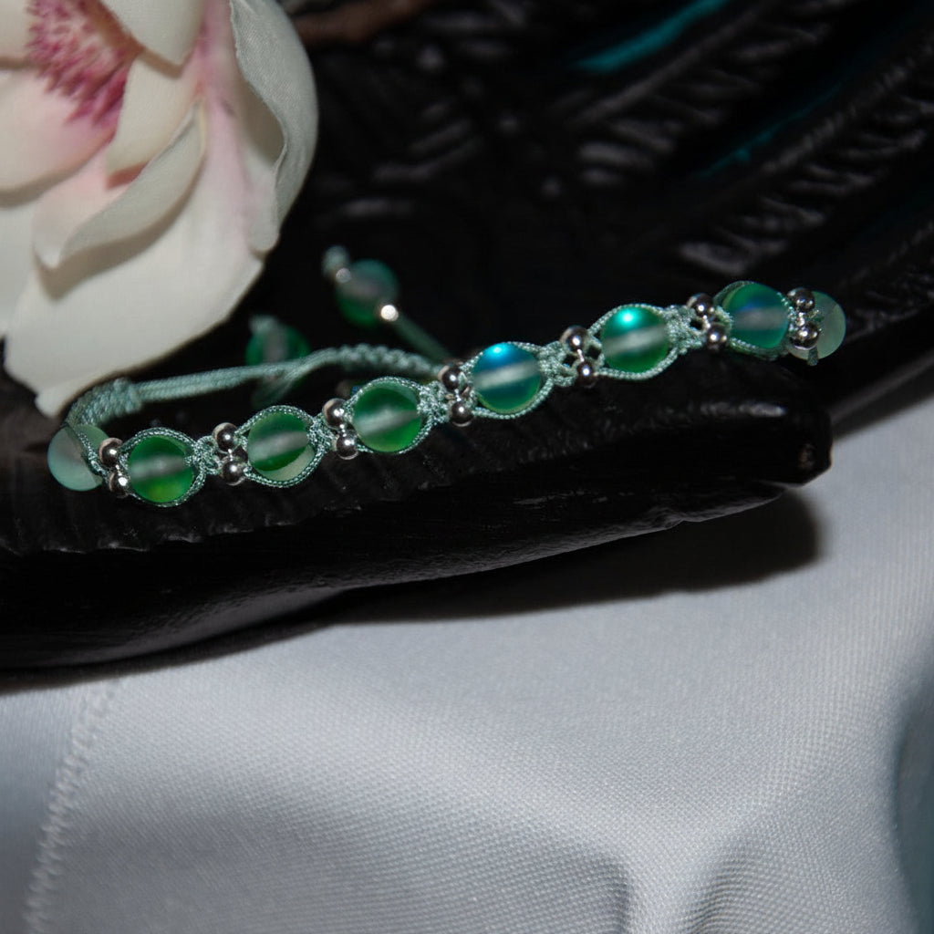 A handmade beaded macrame bracelet with green frosted round beads and silver-toned metal accents, resting on a black leather surface next to a white flower. 