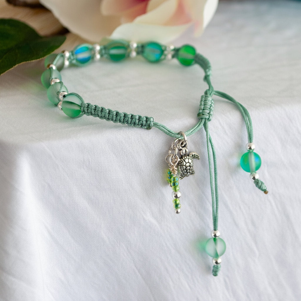 A handmade sage green macrame bracelet with frosted green beads, a small silver turtle charm, and dangling cord beads, displayed on a white fabric surface.