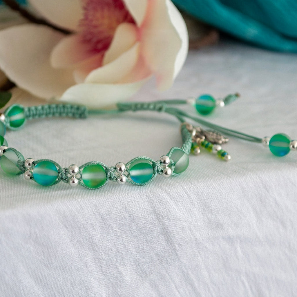 A handmade beaded macrame bracelet with frosted green round beads, silver-toned metal spacer beads, and a small dangling charm, displayed on a white fabric surface with a blurred white flower in the background. 
