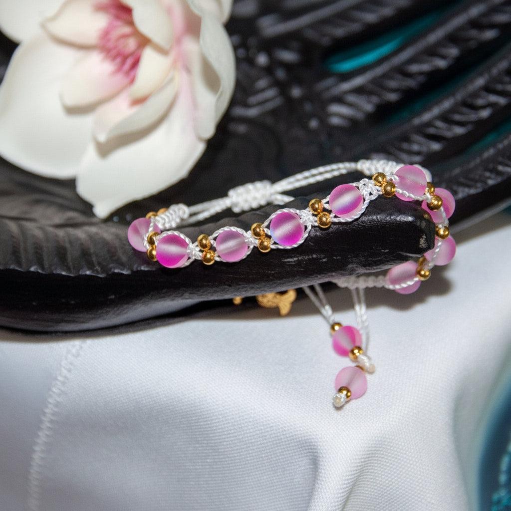 A handmade beaded bracelet with pink frosted glass beads, white cord, and small b spacer rass beads, displayed on a dark textured surface with a white orchid flower in the background. 