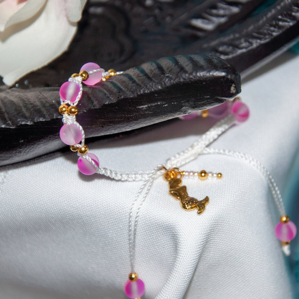 A beaded macrame bracelet with pink frosted beads, white cord, and brass  accent beads, including a small brass mermaid charm, displayed on a white fabric surface with a dark carved prop.