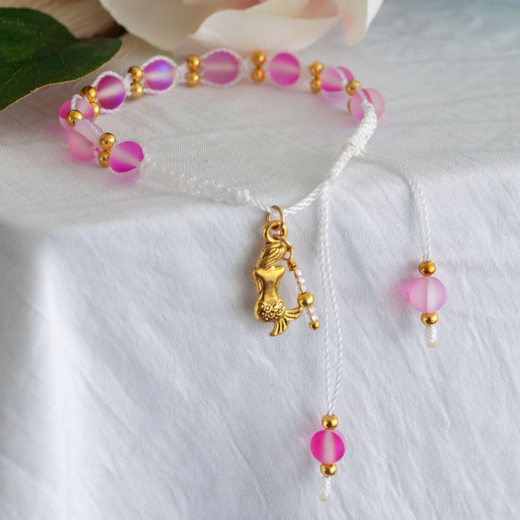 A handmade beaded macrame bracelet with pink frosted round glass beads, brass spacer beads, and a central brass mermaid charm on a white cord, displayed on a white fabric background.