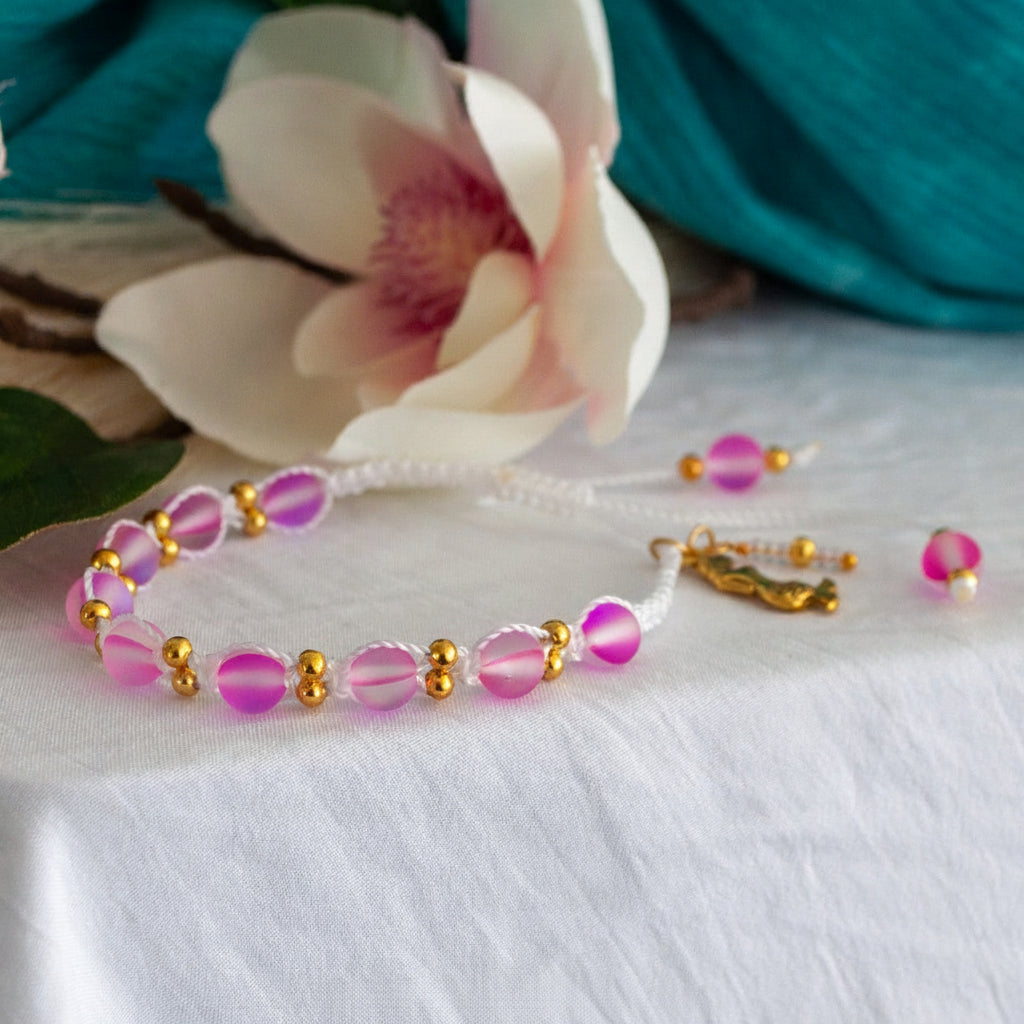 A handmade beaded macrame bracelet with frosted pink round beads, brass spacer beads, and a white cord clasp, displayed on a white fabric surface with a white magnolia flower and teal fabric in the background.