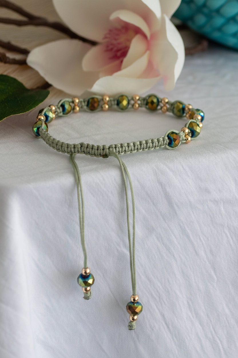 A handmade macrame bracelet in sage green nylon macrame cord with 12 faceted green AB crystal rondelle beads and brass accent beads, displayed flat on white linen showing the full adjustable slide closure with crystal-tipped cord ends and a soft-focus pink magnolia flower in the background