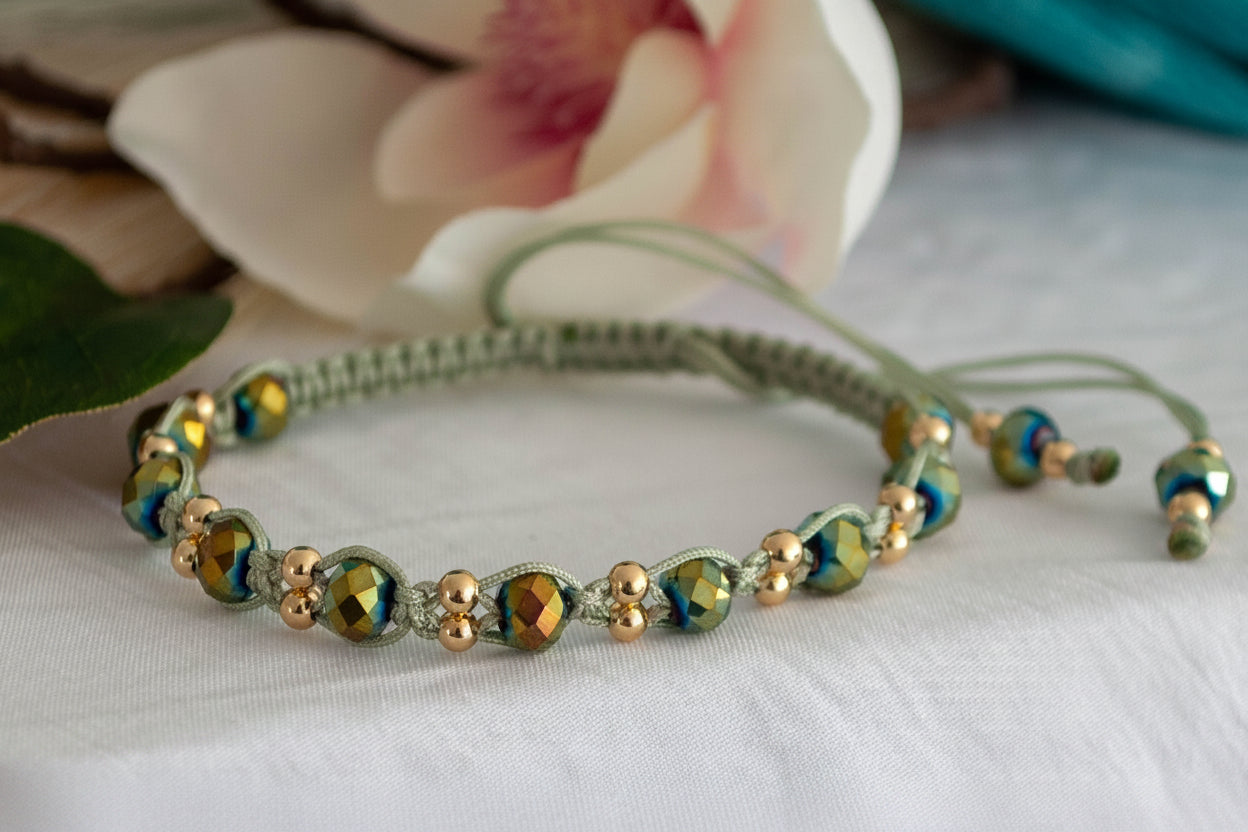 A handmade macrame bracelet in sage green nylon macrame cord with 12 faceted green AB crystal rondelle beads flashing green-gold and small brass accent beads, displayed on a white linen surface with a soft-focus pink magnolia flower and teal background
