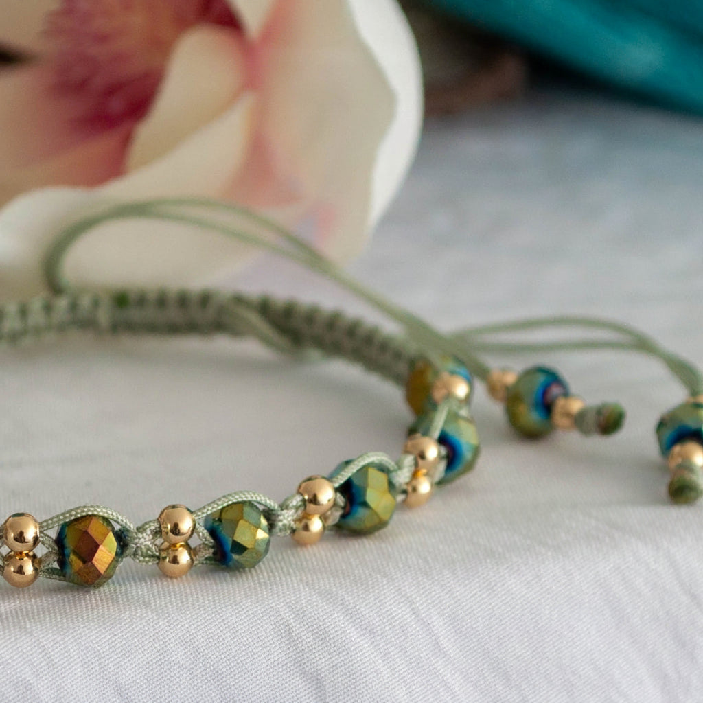 A close-up detail of a handmade macrame bracelet in sage green nylon macrame cord showing faceted green AB crystal rondelle beads with an iridescent green-gold flash and small brass accent beads woven between each crystal, with a soft-focus pink magnolia flower in the background