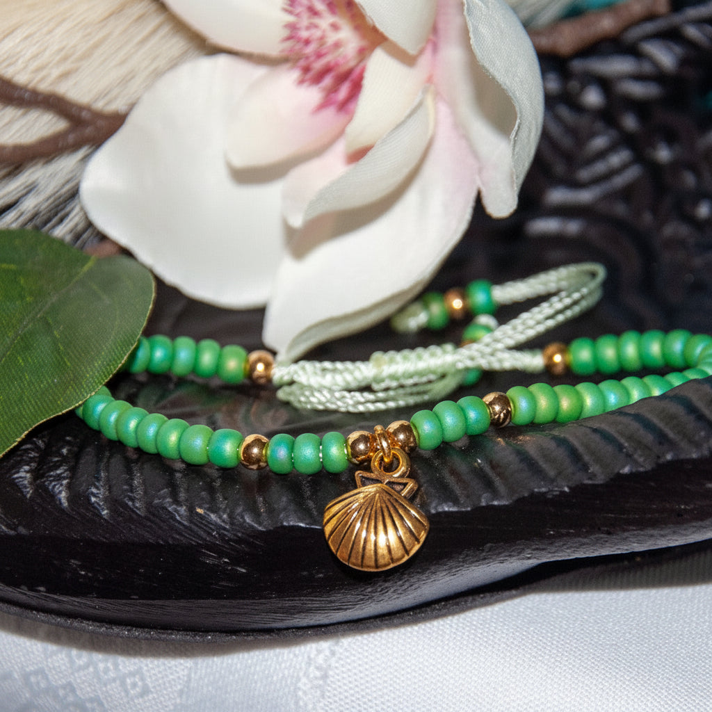 Green beaded bracelet with light green cord and a brass shell charm, displayed on a dark textured tray with a white orchid flower in the background. 