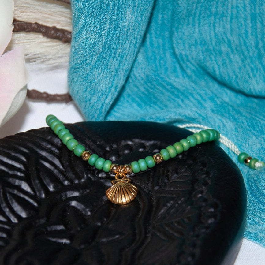 A beaded bracelet with light green round beads, small brass spacer beads, and a brass shell charm, displayed on a black carved stone tray with a turquoise cloth and white flower in the background.