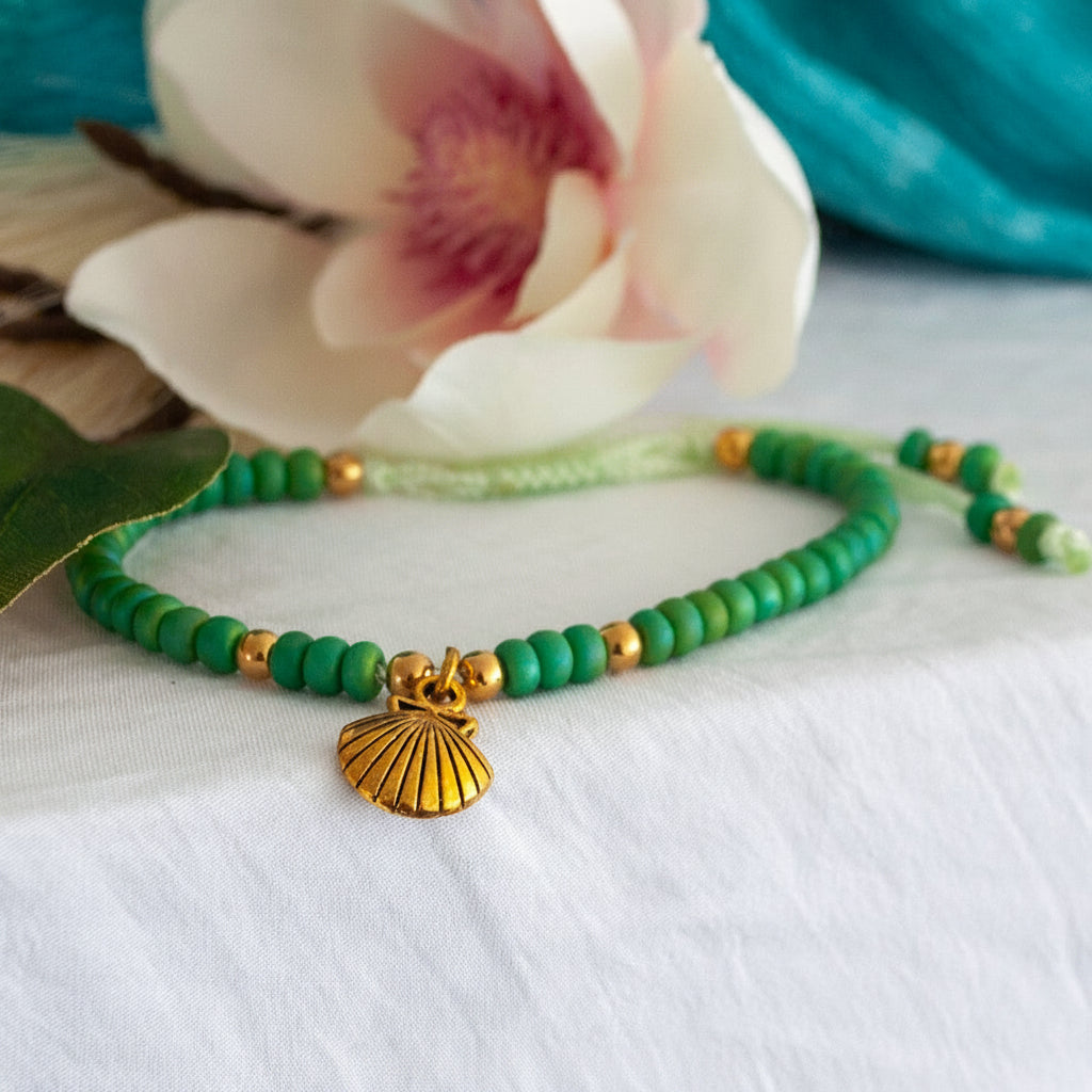 A green beaded bracelet with small brass spacer beads and a brass shell charm, displayed on a white fabric surface with a white flower and teal fabric in the background. 