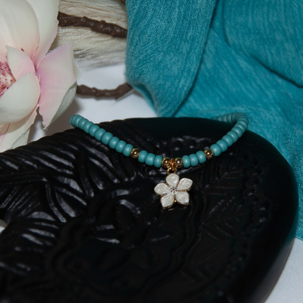 A beaded bracelet with turquoise blue seed beads, small brass spacer beads, and a white enamel flower charm, displayed on a black carved surface with a turquoise fabric and pink flower in the background.