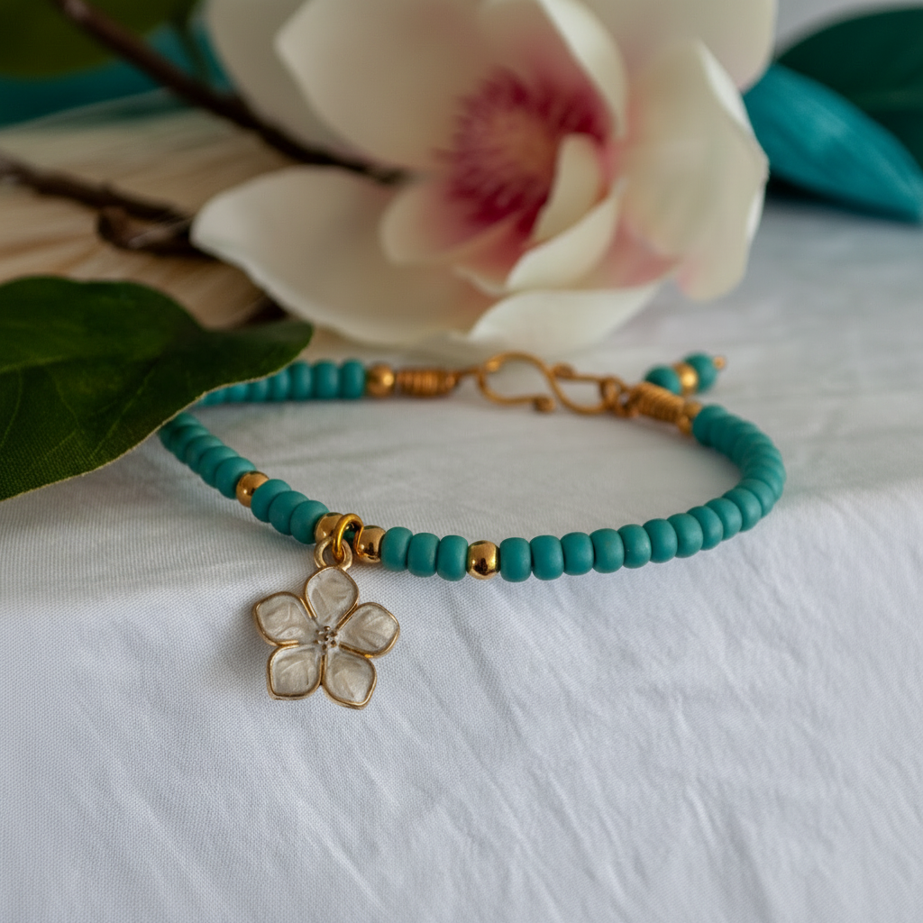 A turquoise blue beaded bracelet with small brass spacer beads and a white enamel flower charm, displayed on a white fabric surface with a blurred flower in the background.