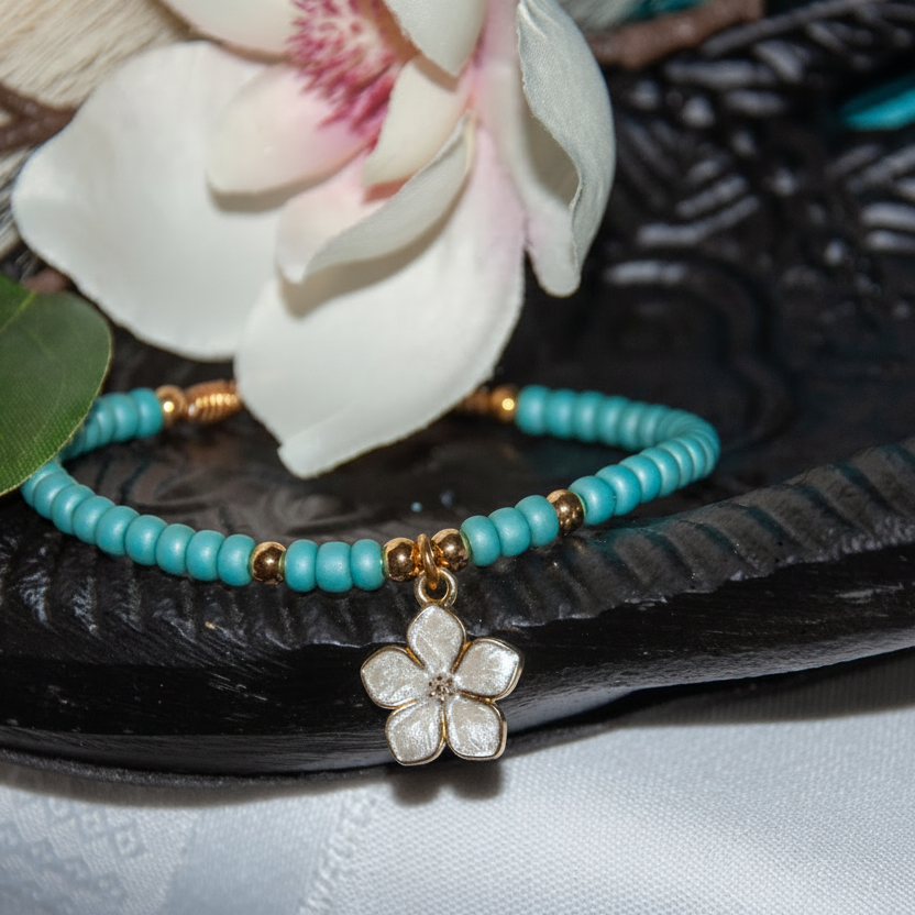 A turquoise blue beaded bracelet with small brass spacer beads and a white enamel flower charm, displayed on a dark carved tray with a white flower prop.