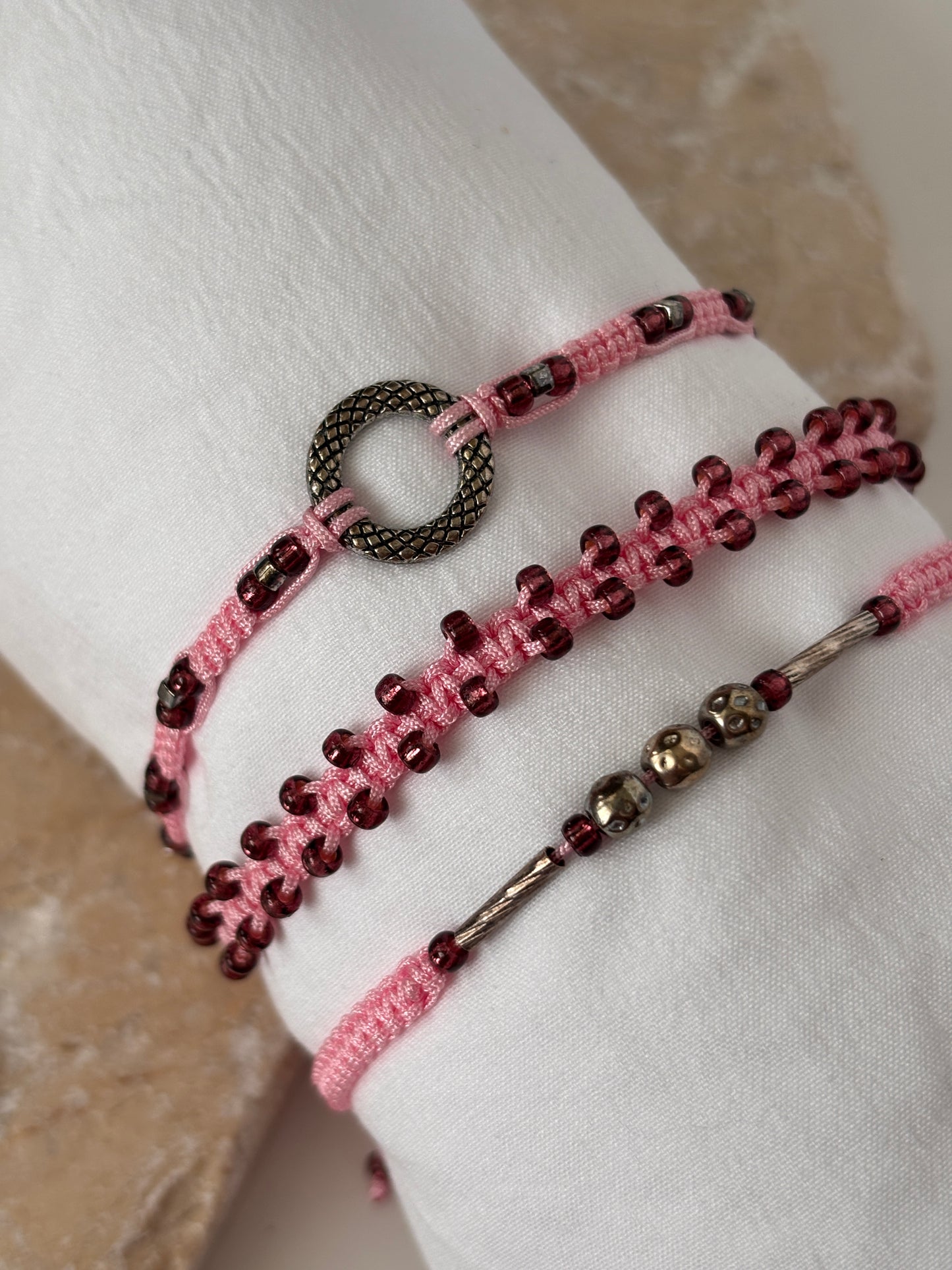A close-up of three pink macrame bracelets on a white linen wrist form against a stone background showing detail of the cranberry seed beads, silver-tone open ring centerpiece, and silver-tone tube and ball accent beads on pink nylon cord.