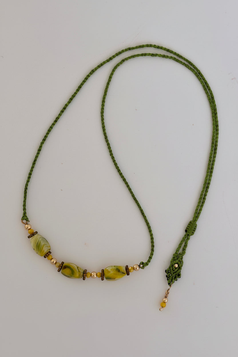 Handcrafted grass green macrame cord necklace with three twisted rectangle chartreuse lampwork glass beads (8mm × 13mm), 4mm round gold-tone beads, 3mm opaque yellow crystal accents, brass rondelle spacers, decorative slide closure, and gold-tone bead dangle, displayed on gray background.