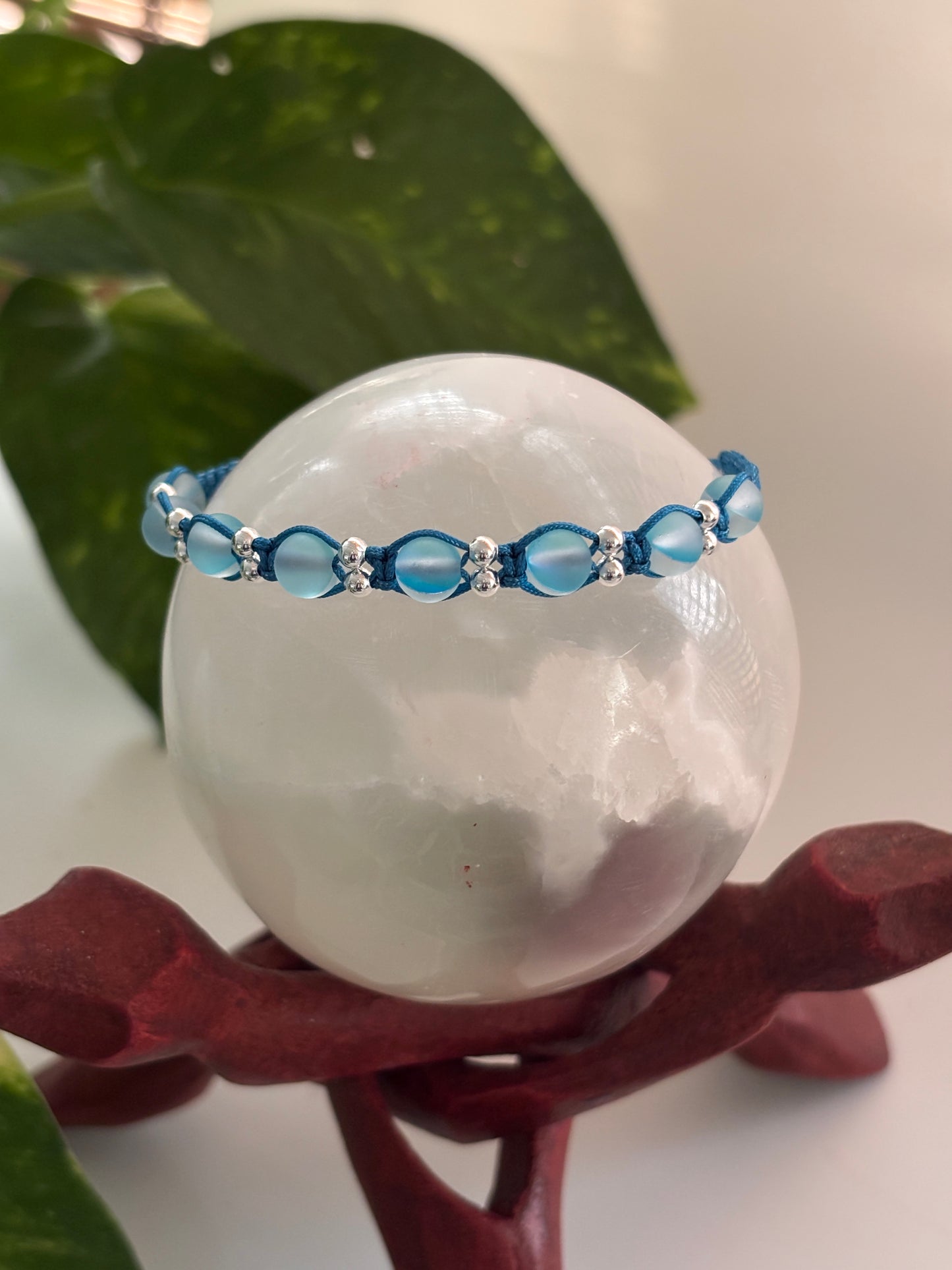 A teal blue beaded macrame bracelet with teal blue frosted mermaid glass beads and silver-tone spacer beads, displayed around a large white crystal sphere on a dark red stand with green leaves in the background.