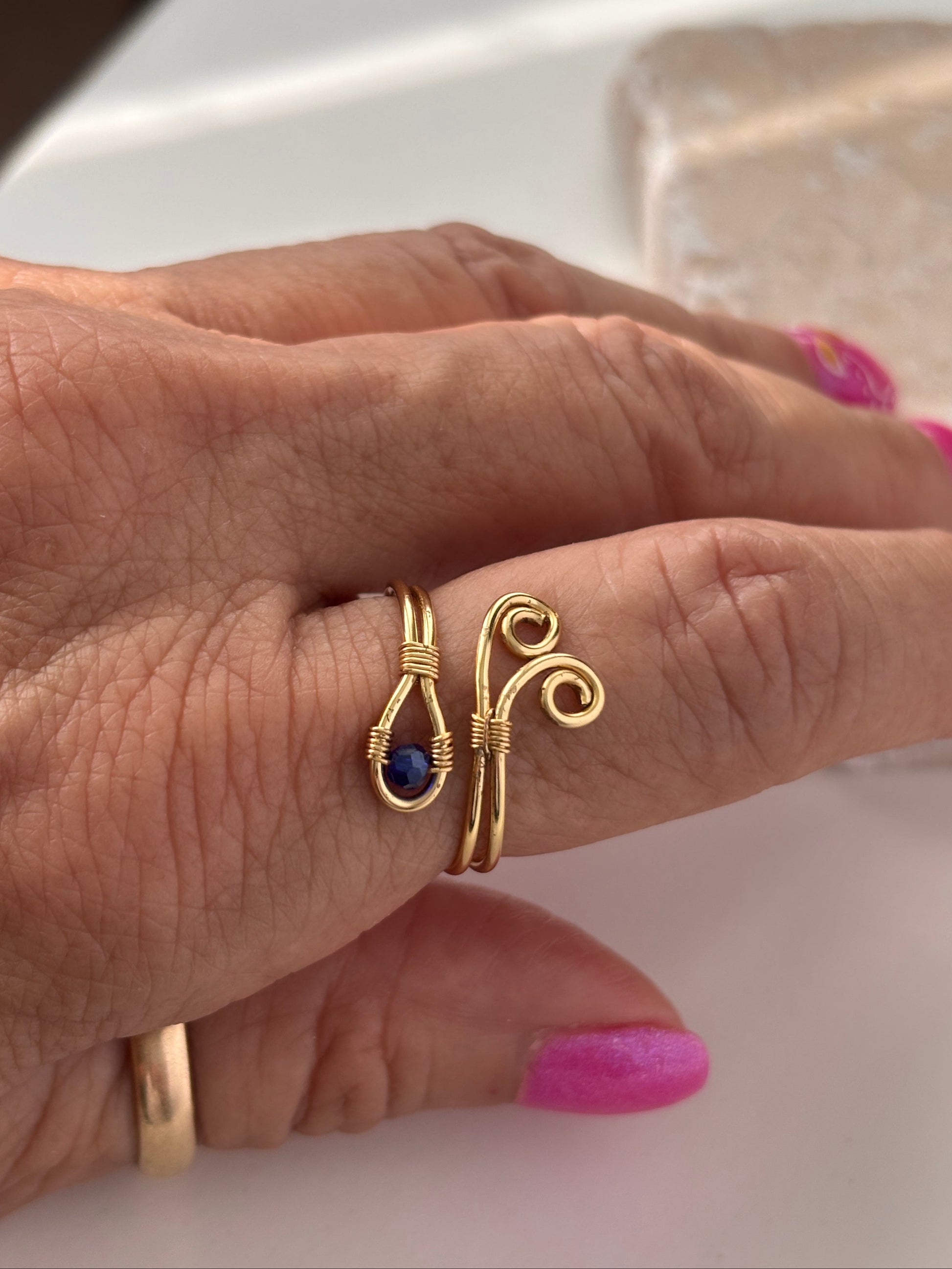 A side view of a handcrafted gold-tone wire ring with a double spiral flourish and a 3mm deep blue faceted crystal bead, worn on a finger resting on a white surface in natural sunlight, with pink nail art in the background