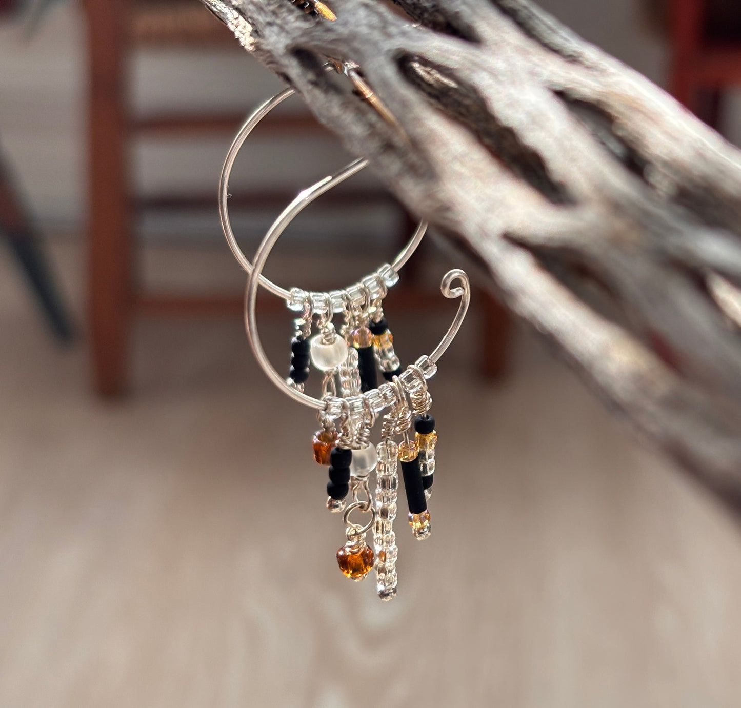A close-up of a pair of handcrafted silver-tone hoop earrings with a scroll-tip wire and five wire-wrapped dangle strands of black bugle beads, clear seed beads, frosted white disc beads, and an amber faceted cube bead drop, hanging from weathered driftwood with a soft-focus wood background.