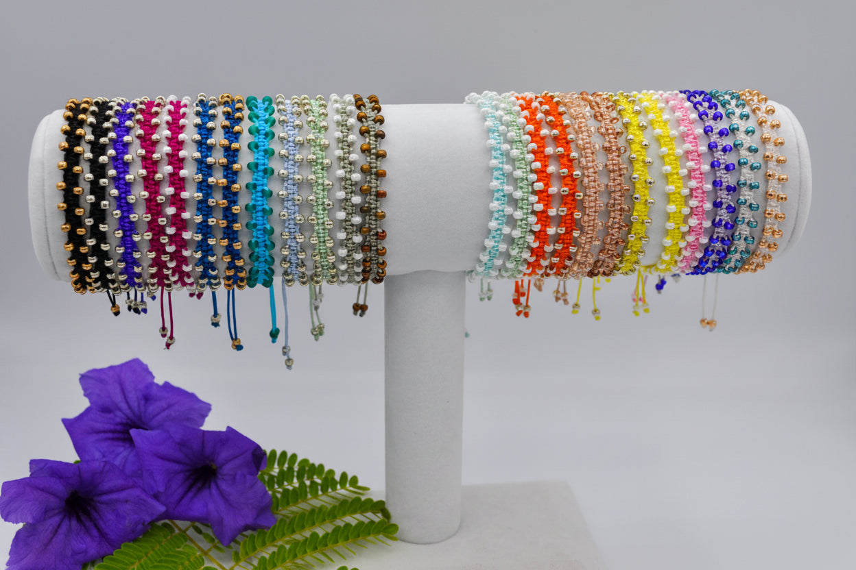 A set of colorful beaded macrame bracelets displayed on a white cylindrical stand, with purple flowers and green leaves in the lower left corner. The bracelets feature various bead patterns and colors including turquoise, silver, gold, orange, yellow, pink, and blue and light peach.