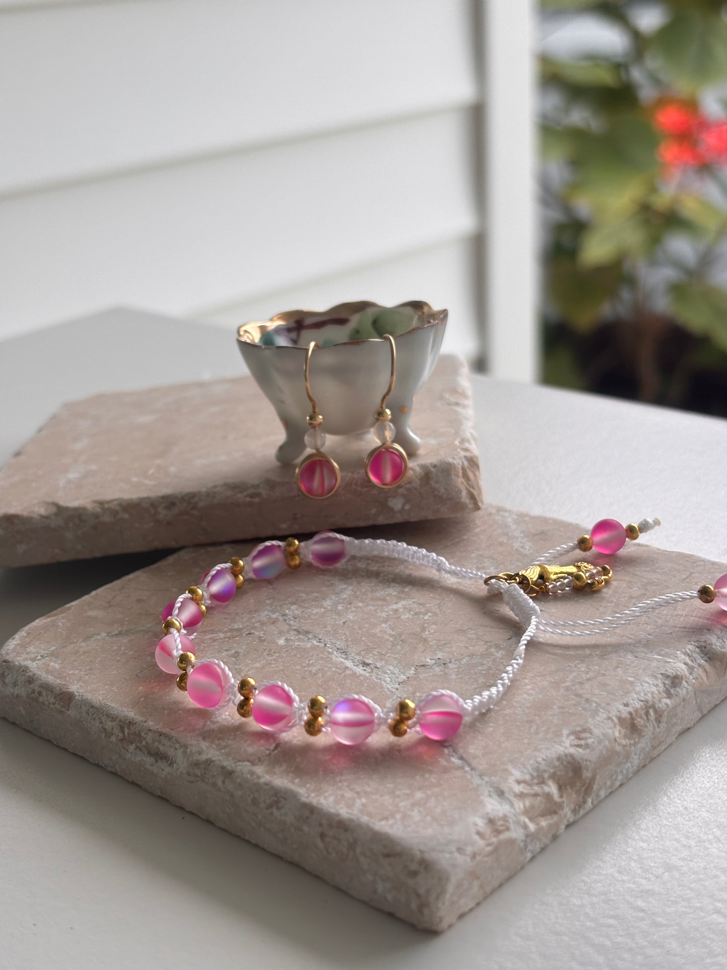 A vivid fuchsia beaded jewelry set including a bracelet and matching earrings with brass accents and white faceted agate beads, displayed on stone slabs with a small silver bowl in the background.  Each item sold separately.