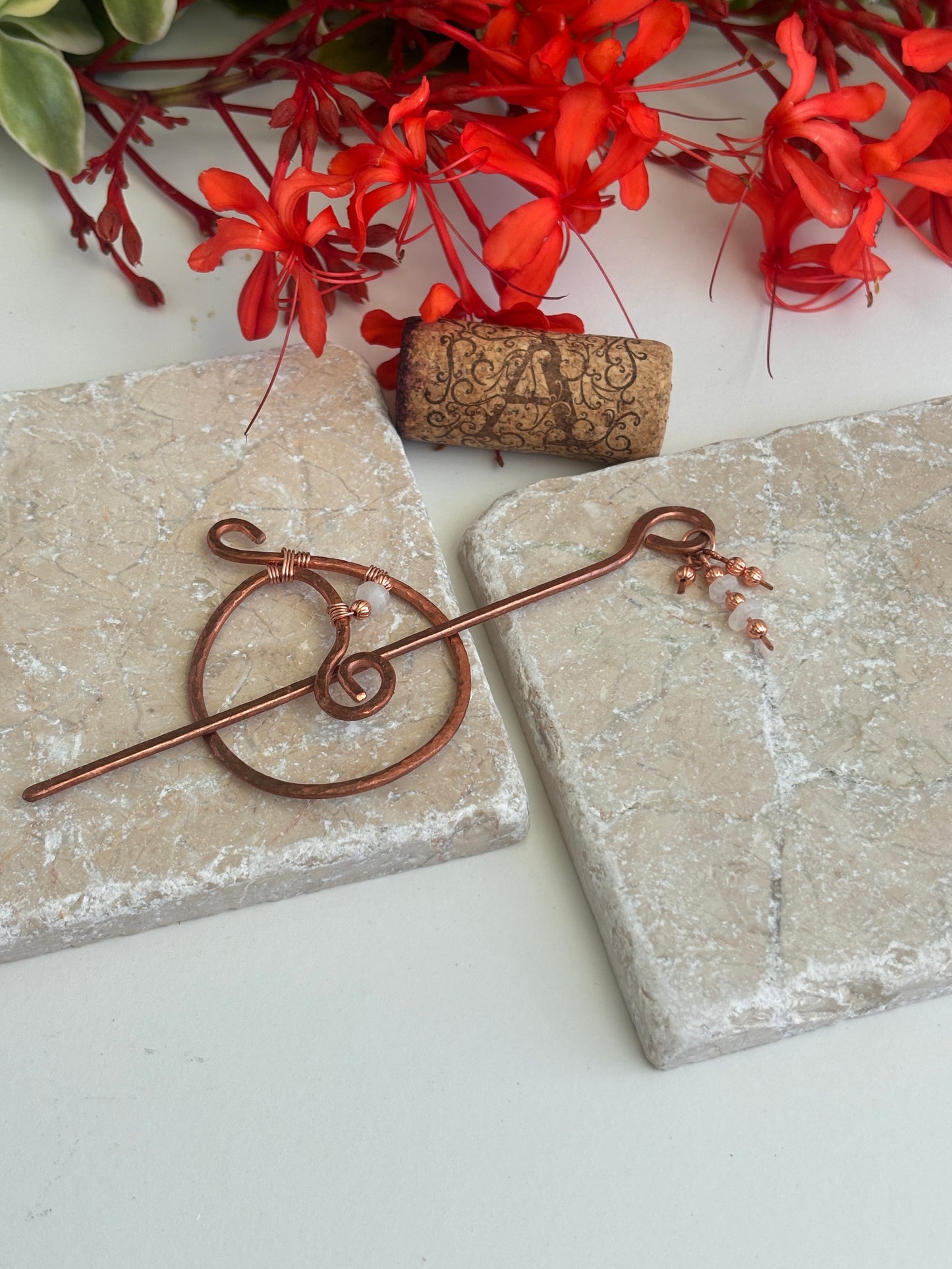 A hammered copper hair pin with a spiral loop and small copper beads  and moonstone beads, styled on two stone tiles with red flowers and a cork in the background.