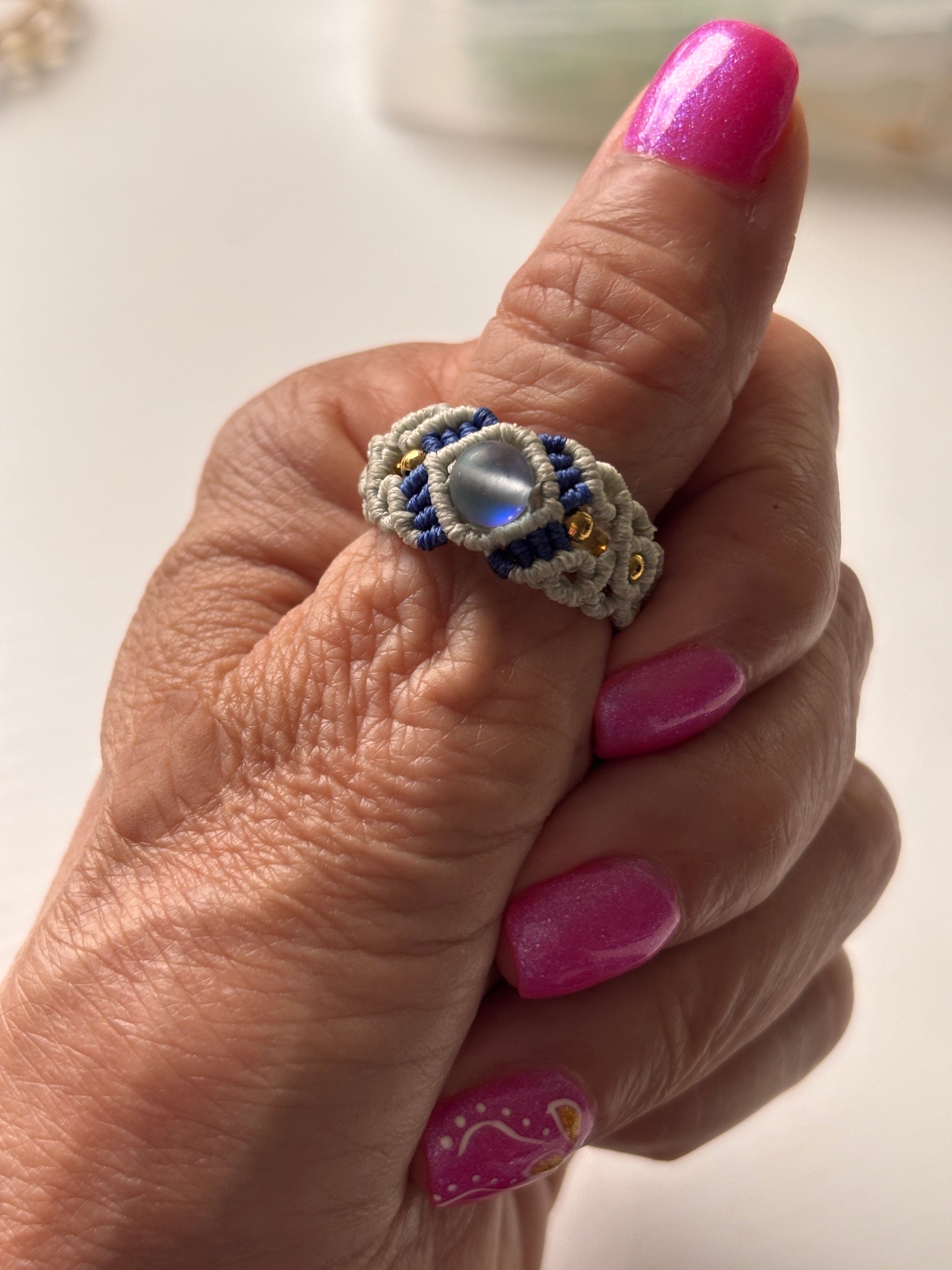 A handmade two-tone light gray and navy blue macrame cigar band ring with a shimmering iridescent mermaid glass bead and gold brass accent beads, worn on a finger with pink nails showing the fit and scale of the ring