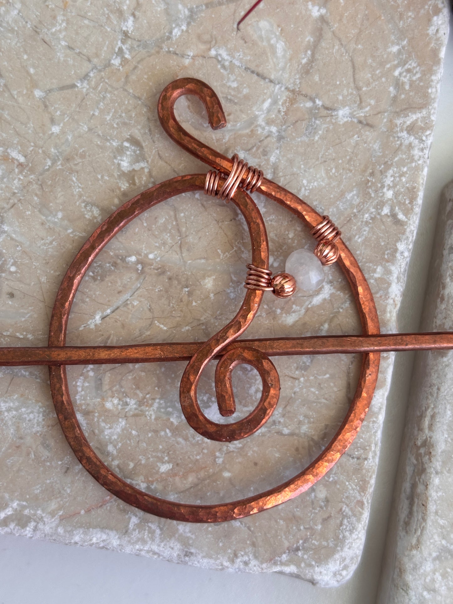 A hammered copper hair stick with a stylized spiral and loop design, accented with copper beads and a moonstone bead, wrapped with wire at key points, displayed on a textured beige stone surface.